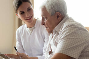 Senior man patient and young woman caregiver medical worker in uniform hold clipboard noting personal information talking listens client telling about health complaints, care support nursing concept