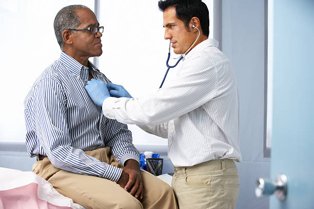 Doctor In Surgery Listening To Male Patient's Chest Using A Stethoscope.