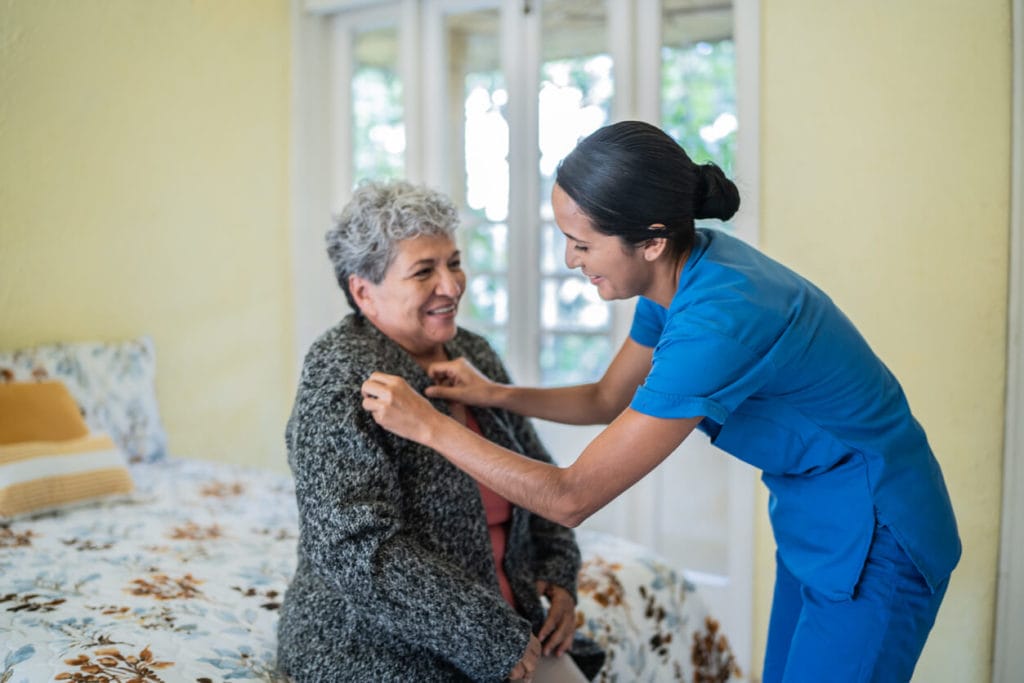 Mid adult nurse helping senior woman dress shirt in bedroom at nursing home