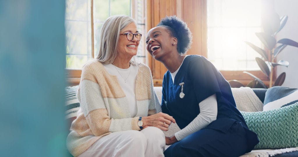 A happy female senior patient is conversing with her female caregiver during healthcare support at the nursing home clinic.